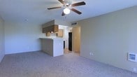 This single-story space is designed around an open-concept layout that connects the living area and kitchen in one cohesive environment. The living area is finished with wall-to-wall carpeting and neutral-toned walls, creating a clean and understated backdrop. A ceiling fan with an integrated light fixture is centrally mounted on the textured ceiling, providing both air circulation and ambient lighting throughout the shared space.

The kitchen is positioned adjacent to the living area and is anchored by a white breakfast bar peninsula that doubles as a casual dining or workspace surface. Upper cabinetry in a warm wood tone is installed above the kitchen counter, offering practical storage. A black refrigerator is visible within the kitchen, and the flooring transitions from carpet to a hard surface in this area, delineating the two zones. A range hood is mounted above the cooking area, reinforcing the kitchen's functional design.

Beyond the kitchen, a hallway leads to additional areas of the unit, with a visible HVAC vent indicating climate control infrastructure. The overall ambiance is practical and straightforward, suited for everyday living with an efficient, open floor plan.