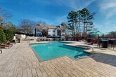 Located at 3209 Westbury Lake Dr in Charlotte, NC, this outdoor amenity space showcases a beautifully appointed resort-style pool area designed for relaxation and recreation. The centerpiece is a generously sized, rectangular swimming pool with crystal-clear turquoise water, accessible via stainless steel entry rails and surrounded by an expansive paver stone deck. The deck provides ample room for lounging, furnished with numerous chaise lounge chairs and shaded by vibrant teal market umbrellas with accompanying seating. A raised terrace area along one side of the pool offers additional seating and is framed by neatly manicured hedgerows, enhancing the sense of privacy and greenery. The space is enclosed by a wrought iron fence, maintaining an open yet secure atmosphere. Towering pine and deciduous trees form a natural wooded backdrop, lending a serene, secluded ambiance to the entire area. A charming white-sided clubhouse building with large multi-pane windows, a prominent chimney, and classic architectural detailing overlooks the pool, anchoring the space with an elegant residential character. The entire outdoor area benefits from abundant natural sunlight, making it a bright and inviting retreat within the community.