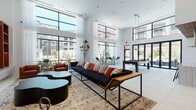 Located at 22121 Clarendon St in Woodland Hills, Los Angeles, this single-floor club room is a thoughtfully designed communal space that blends modern aesthetics with comfortable, functional living. The room is anchored by dramatic double-height ceilings and expansive floor-to-ceiling windows that flood the interior with natural light and offer open views of the surrounding courtyard and residential complex beyond.

The main lounge area features generous seating arranged around a sleek fireplace framed by warm wood built-in shelving, with a mounted flat-screen television serving as a central focal point. Bold artwork and layered area rugs add visual depth and character throughout the space. A billiard table provides an additional recreational element, reinforcing the club room's social atmosphere.

A dedicated dining area accommodates a large communal table beneath a sculptural circular pendant light, paired with a full banquette and curated seating — ideal for gatherings and events. An adjacent full kitchen showcases striking veined marble countertops, a center island with a built-in cooktop, crisp white cabinetry, a textured woven ceiling detail, and stainless steel appliances, making it well-equipped for entertaining. Light tile flooring flows consistently throughout, tying the various zones together into a cohesive, sophisticated environment.