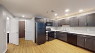 This single-floor unit in Lexington, Kentucky, offers a thoughtfully designed layout with functional living spaces throughout. The kitchen is a standout feature, outfitted with rich dark espresso cabinetry, white countertops, and a full suite of black appliances including a refrigerator, range, microwave, and dishwasher. Recessed lighting illuminates the space, and light wood-look flooring adds warmth throughout the kitchen and adjoining living areas. Additional cabinetry with a double sink provides generous storage and prep space.

The bathroom is designed with accessibility in mind, featuring stainless steel grab bars near the toilet and a spacious layout with light gray walls, wood-look flooring, a large vanity mirror, and dark cabinetry beneath the sink. The walk-in shower area is visible and similarly appointed.

The bedroom offers a comfortable, carpeted floor, soft gray walls, a ceiling fan with integrated lighting, and a window fitted with horizontal blinds that allows natural light to filter into the room.

The overall floor plan, visible in the 3D overview, reveals a cohesive open layout connecting the kitchen and living areas, with the bedroom and bathroom privately situated. The unit's neutral palette and modern finishes create a clean, practical living environment.