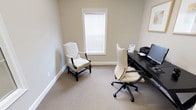 This single-floor office space is designed with clean lines and a neutral palette, creating a calm and professional atmosphere throughout. The walls are finished in a soft warm beige, complemented by crisp white baseboards and trim, giving the space a polished and cohesive look.

The primary workspace is a well-appointed private office featuring a dark espresso L-shaped desk equipped with a computer monitor, printer, and workspace essentials. A high-back cream upholstered office chair sits at the desk, while a classic patterned accent chair offers a secondary seating option for guests or consultations. Natural light filters in through a window fitted with white horizontal blinds, and a pair of gold-framed botanical art pieces adds a refined decorative touch above the workstation. Recessed and overhead lighting ensures consistent illumination throughout the day.

A transitional entryway area connects the main access point to the office interior. This corridor-style space is neatly appointed with a small decorative plant, adding a touch of warmth. Glass-paneled French-style doors with gold hardware divide the entry from the adjoining tiled area, providing both visual openness and a sense of distinction between zones. The consistent use of neutral carpet flooring and light tile ties the space together seamlessly, making it a functional and inviting professional environment.