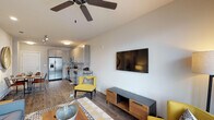 Located at 7740 Midtown Market Avenue in Raleigh, NC, this single-floor unit presents a thoughtfully designed open-concept layout with modern finishes and a cohesive contemporary aesthetic throughout. Wood-look flooring flows consistently across the living, kitchen, and bedroom areas, creating visual continuity and warmth.

The living area features a ceiling fan, a wall-mounted flat-screen television above a mixed-material media console, and natural light from a window fitted with blinds. The space accommodates comfortable seating and offers a relaxed, functional atmosphere.

The kitchen is efficiently laid out along a single wall and is equipped with a full suite of stainless steel appliances, including a French door refrigerator, electric range, built-in microwave, and dishwasher. Light gray flat-front cabinetry extends to the ceiling, complemented by a classic subway tile backsplash and granite-style countertops. Track and flush-mount lighting illuminate the space effectively. A dining area sits adjacent to the kitchen near the entry.

The bedroom is a well-proportioned, serene retreat with a window admitting natural light and a dedicated walk-in closet with built-in shelving. The bathroom features a granite-topped vanity with gray cabinetry, a large wall mirror, modern light bar, tub with shower, and wood-look flooring throughout.