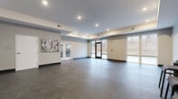 Located at 857A Federal Rd in Brookfield, Connecticut, this single-floor multipurpose space offers a clean, modern design well-suited for fitness and group activities. The open-plan layout provides generous room to move, accommodating a variety of uses from exercise classes to community gatherings.

The interior is finished with durable speckled aggregate flooring throughout, offering a polished yet practical surface ideal for active use. Neutral gray walls are complemented by a distinctive tray ceiling detail with recessed lighting, creating a bright and well-illuminated environment. Dark base molding provides a crisp contrast and grounds the overall aesthetic.

Large floor-to-ceiling windows and glass doors along one wall flood the space with natural light while offering views of the surrounding wooded landscape. A set of white-framed French doors connects to an adjacent fitness room, visible with cardio equipment, allowing for seamless flow between activity areas. Additional standard doors provide access to further amenities within the building.

Decorative wall art adds a touch of warmth to the otherwise contemporary interior. Stackable chairs and folding tables stored along the perimeter indicate the space's flexible, multi-use character. The overall ambiance is clean, open, and functional — a well-appointed amenity space designed to support an active lifestyle.