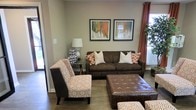 This single-floor space exudes a welcoming and well-curated ambiance, anchored by a thoughtfully designed living area. The living room showcases rich hardwood-style flooring that complements the warm, neutral tones of the walls, creating a cohesive and grounded aesthetic throughout.

The seating arrangement centers around a substantial dark brown sofa accented with a mix of white and burnt orange throw pillows, paired with patterned accent chairs featuring a geometric design in complementary earth tones. A tufted brown leather ottoman serves as a functional and stylish focal point at the center of the arrangement. A sleek end table with a classic table lamp adds soft ambient lighting to the space.

Natural light filters in through windows dressed with bold terracotta-colored curtains, adding a vibrant pop of color that ties the room's warm palette together. Two framed pieces of abstract wall art in shades of orange, green, and blue are displayed prominently above the sofa, lending an artistic character to the room.

A lush potted plant in the corner brings a touch of nature indoors, enhancing the space's inviting atmosphere. Overall, the living area presents a harmonious blend of comfort, style, and warmth, making it a functional and visually appealing gathering space.