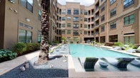 This beautifully designed courtyard amenity space is nestled within the interior of a multi-story residential building, creating a private, resort-like retreat fully enclosed by the surrounding structure. At the heart of the space is a generously sized swimming pool with clear blue water, flanked by a row of modern lounge chairs with vibrant green cushions, ideal for relaxing in the sun. The pool deck is finished with tasteful paver stonework that extends throughout the courtyard, lending a polished, cohesive aesthetic.

Tall palm trees and lush tropical plantings — including broad-leafed cannas and ornamental shrubs — are thoughtfully integrated throughout, adding a verdant, resort-inspired ambiance. Decorative gravel beds accent the base of the palms, contributing textural contrast to the hardscape.

An outdoor kitchen station equipped with a full-size stainless steel grill and granite countertop provides a dedicated space for al fresco cooking and entertaining. Nearby, a paved dining area features outdoor tables and chairs, offering a comfortable setting for gathering. Secured gate access points are visible, ensuring privacy and safety within the courtyard. The multi-story façade surrounding the space features large windows and private balconies, all overlooking this central outdoor retreat.