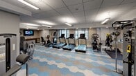 This fitness center is a thoughtfully designed, single-level space offering a comprehensive workout environment. The room is finished with a striking hexagonal-patterned carpet in shades of blue, beige, and white, lending the space a modern and polished aesthetic that distinguishes it from a typical gym setting.

The cardio zone lines one side of the room and features a full row of treadmills, elliptical trainers, upright and recumbent stationary bikes, and stair climbers, all positioned to face windows that provide natural light and outdoor views. A wall-mounted television is also present for entertainment during workouts.

The strength training area is well-appointed with multiple Cybex cable machines, a functional trainer, and various weight-training stations, offering a range of resistance-based exercises. A dumbbell rack and additional selectorized machines occupy additional floor space, accommodating diverse training needs.

The space is finished with a drop-panel ceiling fitted with recessed and pendant fluorescent lighting, ensuring consistent illumination throughout. Roller shades on the windows allow for light control. Entry is through a set of glass-panel double doors, and the neutral gray walls contribute to the clean, professional atmosphere of the facility.