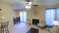 This single-floor space offers a comfortable and functional layout with a seamlessly connected living and dining area. The living room serves as the centerpiece, anchored by a classic white-trimmed fireplace that adds warmth and character to the room. Soft, neutral-toned carpeting runs throughout, complementing the warm beige walls and creating an inviting atmosphere. A ceiling fan with an integrated light fixture is centrally mounted, providing both airflow and ambient lighting.

Natural light filters into the space through slatted window blinds, including a set of French doors that brighten the room and offer access to the exterior. Decorative wall art above the fireplace adds a stylish touch, enhancing the overall aesthetic. The living area provides ample space for seating arrangements, as evidenced by the floral accent chairs flanking the fireplace.

Adjacent to the living room, the dining area accommodates a dark-wood table and chair set, positioned near the French doors to take advantage of the natural light. The open-concept flow between these two areas makes the space feel expansive and well-suited for both everyday living and entertaining.