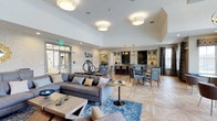 This single-floor clubhouse offers a sophisticated and welcoming environment designed for community gathering and social interaction. The open-concept layout seamlessly connects a comfortable lounge area to a dining and bar space, creating a fluid and inviting atmosphere throughout.

The lounge area is anchored by a plush gray sectional sofa accented with soft throw pillows, complemented by a geometric area rug in blue tones and a sleek low-profile coffee table. The high ceilings, adorned with large drum pendant light fixtures, lend an airy, expansive feel to the space. The floors feature a distinctive light wood herringbone or parquet pattern, adding warmth and visual interest underfoot.

Transitioning deeper into the clubhouse, a dining area with mixed-material tables and blue upholstered chairs offers a refined setting for meals or casual gatherings. Beyond that, a stylishly appointed bar area with dark cabinetry, pendant lighting, and open shelving serves as a natural focal point for social events. Textured wall paneling and tasteful artwork contribute to the space's polished, contemporary character. Large windows allow natural light to filter in, enhancing the bright and welcoming ambiance throughout this well-appointed community clubhouse.