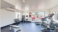 Located in Mesa, Arizona, this dedicated fitness center offers a comprehensive exercise environment designed to support a wide range of workout routines. The space features a clean, modern aesthetic with crisp white walls, recessed lighting, and durable dark flooring that provides both functionality and a professional gym atmosphere.

Natural light flows in generously through multiple large windows positioned along the walls, offering views of the surrounding outdoor area and keeping the space feeling open and inviting. A wall-mounted split air conditioning unit ensures comfortable temperatures year-round, an essential feature in the Arizona climate.

The exercise room is thoughtfully equipped with a variety of cardio machines, including a treadmill, elliptical trainer, and stationary cycle — all Matrix-branded equipment. A multi-functional cable and strength training machine anchors one side of the room, while an adjustable workout bench provides additional versatility for strength exercises.

The free weights section includes a well-organized dumbbell rack stocked with multiple weight increments, complemented by a dedicated storage rack holding medicine balls, stability balls, and a kettlebell for functional training. A wall-mounted flat-screen television adds an entertainment element to workouts. The space is finished with safety equipment including a fire alarm system, making it both practical and compliant.