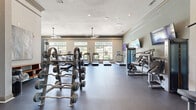 This fitness center offers a spacious, single-floor layout thoughtfully designed to accommodate a full range of workout needs. The open floor plan is finished with durable dark rubber flooring, providing both functionality and a clean, professional aesthetic throughout the space.

The cardio zone lines the windowed wall, where large multi-pane windows flood the area with natural light and provide pleasant views of surrounding greenery. Multiple treadmills, ellipticals, and stair climbers are arranged to take full advantage of these outdoor views. Ceiling fans are suspended overhead, enhancing air circulation during workouts.

The strength training area is equally well-appointed, featuring a cable machine, a variety of selectorized weight machines, benches, and a dedicated free weights section with a full rack of barbells and dumbbells. Multiple wall-mounted flat-screen televisions are positioned throughout the space for entertainment during workouts.

Crown molding and a neutral color palette lend the space an elevated, refined character uncommon in fitness facilities. Glass double doors connect the fitness center to an adjacent common area, adding to the sense of openness. The combination of abundant natural light, quality equipment, and thoughtful design creates a welcoming and motivating environment for users of all fitness levels.