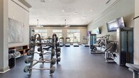 This fitness center offers a spacious, single-floor layout thoughtfully designed to accommodate a full range of workout needs. The open floor plan is finished with durable dark rubber flooring, providing both functionality and a clean, professional aesthetic throughout the space.

The cardio zone lines the windowed wall, where large multi-pane windows flood the area with natural light and provide pleasant views of surrounding greenery. Multiple treadmills, ellipticals, and stair climbers are arranged to take full advantage of these outdoor views. Ceiling fans are suspended overhead, enhancing air circulation during workouts.

The strength training area is equally well-appointed, featuring a cable machine, a variety of selectorized weight machines, benches, and a dedicated free weights section with a full rack of barbells and dumbbells. Multiple wall-mounted flat-screen televisions are positioned throughout the space for entertainment during workouts.

Crown molding and a neutral color palette lend the space an elevated, refined character uncommon in fitness facilities. Glass double doors connect the fitness center to an adjacent common area, adding to the sense of openness. The combination of abundant natural light, quality equipment, and thoughtful design creates a welcoming and motivating environment for users of all fitness levels.
