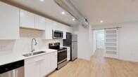 This single-floor unit offers a thoughtfully designed living space that blends modern aesthetics with functional layout. The kitchen serves as a focal point, featuring sleek white cabinetry that extends to the ceiling, providing ample storage. Stainless steel appliances, including a refrigerator, electric range, and built-in microwave, complement the clean, contemporary look. A deep undermount sink with a flexible-neck faucet and light-toned countertops complete the kitchen's polished appearance.

An exposed metallic ventilation duct runs along the ceiling, adding an industrial character that contrasts stylishly with the otherwise clean, minimalist design. Recessed lighting throughout ensures the space is well-illuminated. Light-toned hardwood-style flooring runs continuously through the unit, creating a cohesive and open feel.

A long hallway connects the kitchen and living area to the private spaces, where a sliding barn-style door adds a modern farmhouse touch. The bathroom features frosted glass panel details, enhancing privacy while maintaining a bright, airy atmosphere. The overall design balances industrial and contemporary elements effectively.