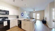 This single-floor residence offers a thoughtfully designed layout with two bedrooms, three bathrooms, a living room, kitchen, hallway, and a closet. The open-concept kitchen and living area serve as the heart of the home, featuring crisp white shaker cabinetry, granite-style countertops, a peninsula with a double-basin sink, and black appliances including a gas range, built-in microwave, dishwasher, and refrigerator. Wood-look flooring flows throughout the main living areas, while pendant lighting and a ceiling fan add both function and style. The living room is bright and airy, benefiting from natural light through a large window and a glass exterior door. The bedrooms offer a calm retreat, each with plush carpeting, ceiling fans, and ample natural light. The primary bedroom provides a comfortable footprint well-suited for a full furniture arrangement. The bathrooms are finished with granite-style vanity tops, white cabinetry, and wood-look flooring. One bathroom features a tiled walk-in shower, while another includes a tub-shower combination with tile surround. A dedicated closet provides additional storage. The overall aesthetic is clean and contemporary, with neutral tones throughout creating a cohesive and inviting atmosphere.