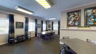 This single-floor event center offers a well-appointed and flexible space designed to accommodate a variety of gatherings, from meetings and social events to private dining. The main area features multiple seating configurations, including a large central conference-style table surrounded by cushioned chairs, ideal for group meetings or communal dining, as well as smaller tables for more intimate settings. The patterned carpet flooring and crown molding trim lend a polished, cohesive aesthetic throughout.

Natural light flows in through multiple windows dressed with white plantation shutters and elegant teal drape panels, creating a bright and welcoming atmosphere. The walls are adorned with bold, colorful framed artwork that adds visual interest and character to the space. Flush and track lighting overhead ensures the interior remains well-lit at all times.

A dedicated kitchenette area features a stone-accented backsplash, counter space, and cabinetry, providing convenient support for catering or event preparation. Adjacent to the kitchenette, a wall-mounted flat-screen television with a media console makes the space equally suited for presentations or entertainment. The layout flows openly between zones, offering adaptability for a wide range of event formats and group sizes.