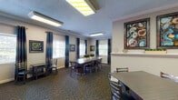 This single-floor event center offers a well-appointed and flexible space designed to accommodate a variety of gatherings, from meetings and social events to private dining. The main area features multiple seating configurations, including a large central conference-style table surrounded by cushioned chairs, ideal for group meetings or communal dining, as well as smaller tables for more intimate settings. The patterned carpet flooring and crown molding trim lend a polished, cohesive aesthetic throughout.

Natural light flows in through multiple windows dressed with white plantation shutters and elegant teal drape panels, creating a bright and welcoming atmosphere. The walls are adorned with bold, colorful framed artwork that adds visual interest and character to the space. Flush and track lighting overhead ensures the interior remains well-lit at all times.

A dedicated kitchenette area features a stone-accented backsplash, counter space, and cabinetry, providing convenient support for catering or event preparation. Adjacent to the kitchenette, a wall-mounted flat-screen television with a media console makes the space equally suited for presentations or entertainment. The layout flows openly between zones, offering adaptability for a wide range of event formats and group sizes.