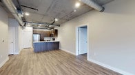 This single-floor space showcases a compelling blend of industrial and contemporary design. The open-plan living and kitchen area is defined by its striking exposed concrete ceiling, which features visible ductwork and utility piping — signature elements of loft-style architecture that lend the space a raw, urban character.

The kitchen is thoughtfully appointed with rich, dark wood cabinetry and a contrasting blue-toned island that serves as both a workspace and a social focal point. Stainless steel appliances, including a full-sized refrigerator, complement the modern aesthetic, while pendant lighting adds warm, focused illumination over the island. A white tile backsplash provides a clean contrast to the darker cabinetry tones.

Warm, light-toned wood-look flooring runs throughout, grounding the industrial elements with a welcoming, residential feel. Recessed lighting supplements the pendants to ensure the space is well-lit throughout. Neutral, light grey walls create a versatile backdrop that allows the architectural details to stand out.

A paneled white door leads to an additional room, suggesting a flexible floor plan. The generous ceiling height and open layout create a strong sense of volume and airiness, making this a well-designed space that balances industrial edge with modern livability.
