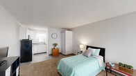 Located at 855 Louisville Rd in Frankfort, Kentucky, this efficient one-bedroom, one-bathroom unit offers a thoughtfully arranged single-floor layout that maximizes every inch of available space. The main living and sleeping area is generously proportioned relative to the overall footprint, featuring wall-to-wall carpeting, neutral gray walls, and a textured ceiling. A large window with vertical blinds allows ample natural light to fill the room, creating a bright and airy atmosphere. The transition from carpet to wood-look vinyl flooring marks the shift into the kitchen and bathroom zones. The kitchen is neatly appointed with white cabinetry, granite-style laminate countertops, a stainless steel sink, and an over-the-range microwave, offering a clean and functional cooking space. The bathroom features a full tub and shower combination with white tile surround, a toilet, wood-look vinyl flooring, and a vanity with a countertop sink and lighted mirror. A curtained partition within the main room provides a flexible division between the sleeping and living areas. The unit's straightforward, open design makes it easy to personalize and maintain, offering a practical living solution in a convenient Frankfort location.
