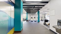 This well-appointed fitness center offers a comprehensive workout environment spread across a single, open floor plan. The space is defined by its bold design language, featuring striking teal-painted structural columns and matching accent walls that contrast sharply against a crisp white and black color scheme. Exposed black ceiling beams run the length of the facility, adding an industrial character while ceiling fans contribute to air circulation throughout.

The cardio zone is well-equipped with multiple treadmills and elliptical machines positioned near large garage-style glass panel windows, flooding the area with natural light and providing outdoor views. A free weights section offers a full dumbbell rack alongside adjustable benches and plyo boxes, catering to a range of strength training needs.

A dedicated functional training area houses a Torque-branded multi-cable rig stocked with medicine balls, kettlebells, battle ropes, and resistance bands. This zone is partially enclosed with glass partitions, offering a defined workout space. Matrix-brand selectorized strength machines occupy a separate section, complemented by athletic-themed wall murals of runners rendered in teal and gold. A water station is conveniently located within the facility. Flooring transitions between carpet and rubberized surfaces to suit each training zone.