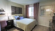 This bedroom is thoughtfully designed to create a calm and restful atmosphere. The room centers around a generously sized bed dressed in soft, neutral-toned bedding, layered with an assortment of decorative pillows in muted blues, greens, and purples. A striking set of dark, textured wall panels serves as a bold headboard backdrop, adding an artistic and grounding focal point to the space. Natural light enters through a window dressed with floor-length mauve curtains hung on grommet rings, which can be drawn to control light and privacy. The walls are painted in a warm, understated tone that complements the overall soothing palette. Plush carpeting in a neutral shade covers the floor, enhancing the room's comfortable and welcoming feel. A dark nightstand positioned beside the bed offers convenient storage and surface space. A framed piece of wall art adorns the adjacent wall, adding a finishing decorative touch. The overall ambiance of the room is serene and well-coordinated, with a cohesive color scheme and layered textures that contribute to a relaxing and inviting environment.