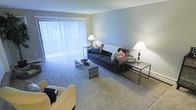 This single-floor residence centers around a well-proportioned living room that serves as the heart of the space. The room is finished with wall-to-wall carpeting in a neutral tone, contributing to a warm and comfortable underfoot feel throughout. Soft gray walls provide a clean, understated backdrop that complements a range of décor styles.

A dominant feature of the living room is the expansive floor-to-ceiling window dressed with vertical blinds, allowing generous amounts of natural light to filter into the space while offering privacy and light control. The window also provides a glimpse of greenery outside, adding a subtle connection to the outdoors.

The layout accommodates a full seating arrangement, including a sofa adorned with colorful accent pillows, flanked by side tables with table lamps that provide warm ambient lighting. A sleek, low-profile coffee table anchors the seating area at the center. Additional seating is visible toward the foreground, suggesting ample room for entertaining or relaxing.

The overall ambiance is clean, functional, and inviting, with a straightforward design that emphasizes comfort and practicality. The space presents a versatile foundation well-suited for a variety of lifestyle needs.