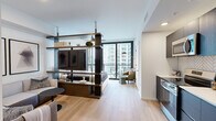 Located in the heart of Atlanta's Midtown neighborhood, this thoughtfully designed studio offers a sleek, modern living experience across a single well-utilized floor. The open-concept kitchen and living area serves as the centerpiece of the space, featuring light hardwood flooring that flows seamlessly throughout. The kitchen is outfitted with dark cabinetry, stainless steel appliances including a range, microwave, dishwasher, and refrigerator, complemented by a striking herringbone-patterned tile backsplash and clean white countertops. A freestanding open shelving unit with dark metal framing acts as a stylish room divider, subtly separating the living and sleeping areas while maintaining an airy, open feel. Floor-to-ceiling sliding glass doors flood the space with natural light and open onto a private balcony, offering sweeping views of the Atlanta skyline. A cozy lounge nook with a sectional sofa provides an intimate retreat within the open layout. The bathroom features large-format light tile flooring, a floating wood-toned vanity with a white countertop, and clean, minimalist finishes. Additional conveniences include an in-unit laundry area and a dedicated closet, rounding out this efficient and stylish urban residence.
