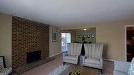 Located at 5014 Sedgewick Dr in Raleigh, NC, this single-story home offers a comfortable and well-appointed living environment with a classic design aesthetic. The living room serves as the heart of the home, anchored by a striking full-height brick fireplace that adds warmth and architectural character to the space. Large vertical blinds along a sliding glass door or window allow for abundant natural light while providing views of the outdoors. The living area is generously sized, accommodating a seating arrangement that includes wingback-style upholstered chairs and a central coffee table, all resting on a patterned area rug over hardwood-style flooring. An open-concept layout connects the living room seamlessly to the dining area, which features an accent wall in a contrasting green tone, decorative wall art, and a dining set beneath a ceiling light fixture. The neutral beige walls throughout create a cohesive and inviting atmosphere, while the textured ceilings add a traditional finish. The overall ambiance of the home is warm and functional, blending classic architectural details like the exposed brick with comfortable, livable spaces suitable for everyday life.
