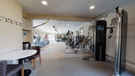 This expansive, single-floor fitness center is designed to accommodate a full spectrum of workout needs in a bright, welcoming environment. The facility is generously proportioned, with an elongated layout that provides ample space between equipment stations, ensuring a comfortable and unobstructed workout experience.

The cardio zone features multiple treadmills with individual screens and a wall-mounted TV, along with elliptical trainers, stationary bikes, and a rowing machine. Large windows throughout this section overlook an outdoor pool area, flooding the space with natural light and offering refreshing views during workouts.

The strength training area is equally well-appointed, housing a full rack of dumbbells, adjustable benches, a barbell rack, and multiple cable and weight machines including Precor and Maxican branded systems. A dedicated stretching and functional fitness zone includes stability balls, medicine balls, and TRX suspension trainers anchored to the ceiling.

Full-length mirrors line key walls, visually expanding the space and supporting proper form during exercise. The flooring features a durable combination of rubber and tile in a neutral checkerboard pattern, and recessed lighting along with ceiling fans maintain a comfortable atmosphere throughout. Built-in shelving and a small seating area complete this comprehensive facility.
