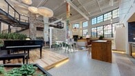 Located at 2048 Fulton Rd in Cleveland, OH, this single-floor space is a dynamic, loft-style community and event venue that masterfully blends industrial architecture with warm, creative design. Soaring ceilings with exposed steel beams, skylights, and an expansive grid of large black-framed windows flood the interior with natural light and offer views of the surrounding neighborhood.

The open floor plan is anchored by a raised hardwood platform stage featuring a grand piano, making it ideal for live performances and gatherings. Polished terrazzo flooring transitions seamlessly into rich hardwood, delineating distinct zones throughout the space. A central gathering area is furnished with eclectic lounge seating, a fireplace, and a built-in wood bar counter, while house-shaped booth enclosures provide intimate nooks for smaller conversations or work.

Throughout the space, bold art installations command attention — from a towering stacked-letter sculpture reading "Home on the Range," to vibrant large-format canvas paintings and sculptural red metalwork. Lush indoor planters add a natural, living quality to the environment. An industrial steel staircase leads to a mezzanine-level overlook, adding vertical dimension. The overall ambiance is energetic, welcoming, and artistically rich.