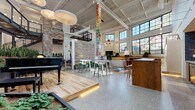 Located at 2048 Fulton Rd in Cleveland, OH, this single-floor space is a dynamic, loft-style community and event venue that masterfully blends industrial architecture with warm, creative design. Soaring ceilings with exposed steel beams, skylights, and an expansive grid of large black-framed windows flood the interior with natural light and offer views of the surrounding neighborhood.

The open floor plan is anchored by a raised hardwood platform stage featuring a grand piano, making it ideal for live performances and gatherings. Polished terrazzo flooring transitions seamlessly into rich hardwood, delineating distinct zones throughout the space. A central gathering area is furnished with eclectic lounge seating, a fireplace, and a built-in wood bar counter, while house-shaped booth enclosures provide intimate nooks for smaller conversations or work.

Throughout the space, bold art installations command attention — from a towering stacked-letter sculpture reading "Home on the Range," to vibrant large-format canvas paintings and sculptural red metalwork. Lush indoor planters add a natural, living quality to the environment. An industrial steel staircase leads to a mezzanine-level overlook, adding vertical dimension. The overall ambiance is energetic, welcoming, and artistically rich.