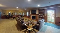Located at 4010 Clearwater Rd in Saint Cloud, Minnesota, this single-floor gathering space offers a rich, traditional aesthetic defined by warm wood wainscoting, carpeted flooring, and classic furnishings throughout. The main lounge area is generously proportioned, anchored by a stone-surround fireplace with a mounted flat-screen television above, and flanked by built-in bookshelves stocked with reading material. Comfortable leather sofas and wrought-iron glass-top coffee tables create an inviting seating arrangement ideal for group gatherings. Adjacent to the lounge, a billiards area features a full-size pool table illuminated by a hanging light fixture, adding a recreational dimension to the space. A foosball table is also present, further enhancing the entertainment options. The fully equipped kitchen includes stainless steel appliances — refrigerator, range, and microwave — rich cherry cabinetry, tile flooring, and a pass-through counter that connects to the main area. A lounge corridor lined with high-top pub tables, leather bar stools, and decorative mirrors provides additional seating with a social atmosphere. Natural light filters in through windows overlooking the outdoors, complementing the warm ambient lighting provided by decorative pendant fixtures throughout.