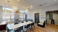 This single-floor community lounge is designed with an open-plan layout that encourages social interaction and relaxed gatherings. The space features warm hardwood flooring throughout, paired with a drop ceiling fitted with recessed lighting and a series of eye-catching, orb-shaped pendant fixtures that serve as decorative focal points. Soaring windows along multiple walls flood the interior with natural light and offer views of the surrounding neighborhood, creating a bright and welcoming atmosphere.

The main area is furnished with a mix of seating arrangements, including large communal tables with modern black chairs for group gatherings, as well as smaller high-top bistro tables and bar stools positioned near the windows for more intimate settings. A comfortable wicker lounge chair is tucked near a floor-to-ceiling glass door that opens to an outdoor patio area. A shuffleboard table adds a recreational element to the space, complemented by wall art that contributes to the room's contemporary character.

A compact kitchenette is tucked to one side of the space, equipped with rich wood cabinetry, granite-style countertops, a stainless steel sink, microwave, and refrigerator. Restroom access is also available within the space. Overall, the lounge offers a functional yet stylish environment suited for community use.