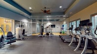 Located at 6650 Corporate Center Pkwy in Jacksonville, FL, this fitness center offers a comprehensive workout environment designed to support a wide range of exercise routines. The facility is organized across a single floor and divided into distinct functional zones, each tailored to specific training needs.

The main cardio area features a row of treadmills, elliptical machines, and stair climbers positioned along a windowed wall, allowing natural light to filter in and providing outdoor views of the surrounding greenery. Mounted televisions are conveniently placed for entertainment during workouts. Ceiling fans complement the recessed lighting overhead, contributing to a comfortable exercise environment.

A strength training section is equipped with cable machines, weight benches, and a variety of resistance equipment. Full-length mirror panels line the walls throughout, visually expanding the space and allowing users to monitor their form. A separate spin cycle area features hardwood flooring and multiple stationary bikes, set apart by glass partition walls that maintain an open feel while defining the zone.

An adjacent room, visible through the glass partitions, houses a basketball court, adding a recreational dimension to the facility. The interior design incorporates teal and warm neutral tones, with rubber flooring in the main workout areas ensuring durability and safety. The overall layout is spacious, well-organized, and suited for diverse fitness activities.