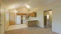 This single-story space showcases an open-concept layout where the kitchen flows seamlessly into the living area. The kitchen is a standout feature, highlighted by soaring vaulted ceilings that lend an airy, spacious feel to the room. Warm honey-toned oak cabinetry lines the walls, offering generous storage, and is complemented by light countertops that extend along the perimeter and across a peninsula breakfast bar — ideal for casual dining or meal prep. The kitchen is equipped with white appliances, including a refrigerator, electric range, and built-in microwave. A double sink is positioned beneath upper cabinetry near the corner. The flooring transitions from easy-care light vinyl in the kitchen to plush carpet in the adjacent living area, creating a comfortable and defined separation between spaces. A chandelier adds a touch of warmth near the dining area, enhancing the overall ambiance. An arched entryway leads to additional areas of the home, hinting at a well-connected floor plan. The neutral color palette throughout gives the space a clean, welcoming atmosphere with strong potential for personalization.