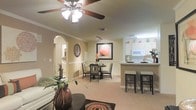 This single-floor residence offers an open-concept layout that effortlessly connects the living, dining, and kitchen areas into one cohesive and functional space. The living area is anchored by a ceiling fan with integrated lighting and is finished with plush carpeting that adds warmth underfoot. Crown molding runs throughout, lending a polished, refined character to the interior. A graceful arched doorway adds architectural charm, leading to additional areas of the home. The dining area accommodates a round table with wicker-style chairs, creating an intimate setting for meals, while bold wall art and decorative accents contribute personality and color to the neutral, warm-toned walls. Adjacent to the dining space, a peninsula-style kitchen features white cabinetry, built-in appliances including a microwave, and a breakfast bar with seating — ideal for casual dining and conversation. Track lighting illuminates the kitchen area efficiently. The cohesive color palette of soft beige and cream tones throughout the space creates a bright, welcoming ambiance. Thoughtfully placed greenery and decorative accessories enhance the overall aesthetic, making this a comfortable and visually appealing place to call home.