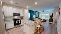 This thoughtfully designed single-floor residence combines modern finishes with a lively, eclectic aesthetic. The open-concept kitchen is a standout feature, outfitted with crisp white shaker cabinetry, granite countertops, stainless steel appliances, and a central island with an integrated sink and bar seating — ideal for casual dining and entertaining. Light wood-look flooring flows seamlessly from the kitchen into the living area, where a bold teal accent wall serves as a dramatic backdrop for a gallery of colorful abstract artwork and a ceiling fan overhead. Natural light filters in through multiple windows dressed with blinds, and a glass door provides direct outdoor access. The bedrooms offer a warm, inviting atmosphere; one features a decorative chandelier, a rattan statement headboard, vibrant patterned rugs, and a window that brings in soft natural light. A second bedroom is bright and airy, with carpet flooring, outdoor access via a glass door, and ample wall space. The bathroom is well-appointed with granite countertops, white cabinetry, a framed glass shower, and wood-look flooring. Throughout the residence, recessed lighting, clean lines, and a cohesive blend of bohemian and contemporary design elements create a dynamic and welcoming living environment.