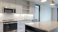 This single-floor unit showcases a thoughtfully designed open layout with consistent modern finishes throughout. The kitchen serves as a central highlight, featuring crisp white shaker-style cabinetry with under-cabinet lighting, granite countertops, and a substantial island with a contrasting steel-blue base topped with matching granite. Stainless steel appliances, including a French door refrigerator, range, and microwave, complement the subway tile backsplash. Gold pendant lighting adds a refined touch above the island.

The living area is anchored by warm hardwood flooring that flows seamlessly throughout the main spaces. A set of bold black-framed French doors floods the room with natural light while providing outdoor access. Bedrooms continue the hardwood flooring theme, with large windows fitted with blinds offering additional natural light. A ceiling fan is present in one bedroom for added comfort.

The walk-in closet offers generous built-in shelving and hanging rods with dark metal hardware. The bathroom features a double-sink vanity with granite countertops, gray cabinetry, a tiled tub-shower combination, and large-format marble-style flooring, creating a clean, spa-inspired atmosphere.