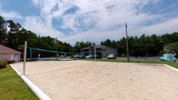 This outdoor amenity space in Pensacola, Florida offers a well-rounded collection of recreational features designed for active outdoor living. The centerpiece sand volleyball court is fully equipped with a regulation net and provides a generous playing surface, set against a backdrop of mature trees and open sky. Nearby, a spacious resort-style swimming pool is surrounded by a wide concrete deck furnished with lounge chairs and shaded by navy blue umbrellas, with tropical palm trees framing the perimeter and a white fence enclosure providing definition to the space.

A covered walkway runs alongside a building exterior finished with stacked stone accents and lap siding, illuminated by classic wall-mounted lanterns. The walkway connects to an outdoor patio area furnished with a table, chairs, and a large umbrella, ideal for casual gathering. French doors with slatted blinds provide access to the interior spaces.

Perhaps the most scenic element is the community pond, featuring a central decorative fountain and bordered by well-maintained green lawns. The reflective water and lush surroundings create a tranquil natural atmosphere. The entire outdoor space benefits from Florida's abundant natural light and open skies, making it a vibrant and versatile amenity area.