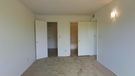 This single-floor space presents a functional bedroom that is designed with convenience in mind. The room is finished with wall-to-wall carpet in a neutral beige tone, contributing to a quiet and comfortable atmosphere. The walls are painted in a clean, off-white shade, lending a simple and understated aesthetic throughout.

A notable feature of this bedroom is its direct access to a bathroom, visible through one of the centrally positioned doorways. The bathroom appears to include a bathtub or shower and additional storage, making it a practical en-suite arrangement. Flanking the bathroom entry are two additional doors — one leading to a spacious walk-in closet equipped with a closet rod for hanging storage, and another that appears to access a secondary closet or utility space.

Overhead, a ceiling vent indicates a central HVAC system for climate control. The room offers a generous sense of space, with enough floor area to accommodate standard bedroom furnishings comfortably. Overall, the space provides a clean, move-in ready environment with an emphasis on storage accessibility and practical functionality.
