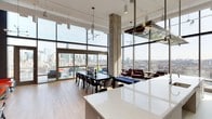 This remarkable single-floor space makes an immediate impression with its dramatic, double-height floor-to-ceiling glass walls that flood the interior with natural light and frame breathtaking panoramic city skyline views. Exposed concrete columns punctuate the open-plan layout, lending an industrial-chic character that is both raw and refined.

The living and dining areas flow seamlessly together, centered around a large dining table comfortably accommodating a generous number of guests, complemented by sleek pendant lighting overhead. The lounge area is furnished with rich, deep-toned upholstered seating, adding warmth and contrast against the cool concrete and glass aesthetic.

The kitchen features a pristine, high-gloss white countertop island that anchors the space, offering both functionality and a clean, contemporary aesthetic. Track lighting and modern pendant fixtures throughout ensure the space is well-lit even after dark.

Light hardwood flooring runs throughout, softening the industrial elements and adding a sense of warmth. The overall ambiance is one of elevated urban sophistication — an open, airy, and thoughtfully designed space that balances modern architecture with comfortable, livable interiors, all while showcasing expansive city views from nearly every vantage point.