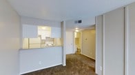 This single-floor space offers a functional, open-concept layout that connects the main living area to the kitchen via a convenient pass-through counter. The kitchen is equipped with white cabinetry, a white electric range, and a refrigerator, with overhead lighting providing a well-lit workspace. The granite-style countertop adds a practical surface along the pass-through, making the kitchen feel connected to the main living space.

The living area features wall-to-wall carpeting in a neutral tone, contributing to a warm and comfortable atmosphere. A textured ceiling runs throughout, and a centrally located HVAC vent ensures climate control across the space.

A hallway extends from the living area, leading to a bathroom and additional rooms beyond. The bathroom is visible from the hallway and includes standard fixtures. The hallway is well-lit by a flush-mount ceiling fixture, providing clear sightlines through the unit. The overall color palette is neutral throughout, with white walls and trim creating a clean, versatile backdrop suitable for a variety of interior styles.