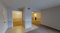 This single-story space showcases an open, interconnected layout that allows for seamless flow between its main living areas. A central structural column serves as a subtle architectural divider, separating the tiled entryway or transitional zone from the carpeted living and dining areas. The flooring shift from light ceramic tile to neutral-toned carpet adds visual distinction between spaces without the use of walls.

To one side, a dining area is highlighted by a classic chandelier that casts warm, inviting light, complementing the room's soft, neutral wall tones. On the opposite side, an opening leads to what appears to be a kitchen, where cabinetry and appliances are visible, suggesting a functional and efficiently arranged cooking space. The arched entryway detail adds a subtle architectural touch.

Throughout the space, consistent light gray walls and white trim create a clean, cohesive aesthetic. HVAC vents are integrated into the ceiling, ensuring climate comfort. Baseboards run uniformly along each room, reinforcing the tidy, well-maintained character of the interior. The overall ambiance is calm, neutral, and move-in ready.