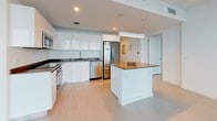 Unit 49F is a contemporary high-rise residence designed with a clean, modern aesthetic and an abundance of natural light. The open-concept living and dining area is defined by dramatic floor-to-ceiling glass walls that frame breathtaking panoramic views of the city skyline and waterfront, flooding the space with sunlight and creating a seamless connection to the outdoors via a private balcony.

The kitchen is outfitted with high-gloss white cabinetry, dark countertops, and a full suite of stainless steel appliances including a French-door refrigerator, built-in oven, cooktop, microwave, and dishwasher. A central island with a warm-toned countertop provides additional workspace and casual seating, anchoring the kitchen within the open living area.

Light wood-tone flooring extends throughout the main living spaces and bedrooms, lending warmth and continuity. The bedrooms each enjoy direct access to the expansive water and city views through large sliding glass doors leading to the balcony.

The bathrooms are finished with striking marble-patterned tile flooring and walls, white vanities with chrome fixtures, and glass-enclosed showers and tub combinations. A generously sized walk-in closet with built-in shelving and hanging rods provides ample storage. A dedicated laundry area with in-unit washer and dryer completes this well-appointed residence.