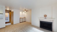 This single-floor space offers a clean, functional layout centered around a welcoming living area. The room is finished with neutral wall-to-wall carpeting and crisp white walls, creating a bright and airy atmosphere throughout. A standout feature is the tile-surround fireplace positioned along one wall, offering a classic focal point that adds warmth and character to the space. Opposite the fireplace, a built-in storage cabinet with wood-framed panel doors provides practical storage while adding a warm, mid-century-inspired design touch. A ceiling fan with integrated lighting is centrally mounted, ensuring comfortable air circulation and ambient illumination. The open layout allows for flexible furniture arrangements, making the living area highly adaptable to various uses. Recessed accent lighting highlights the cabinet area, adding depth and dimension to the room's overall aesthetic. The combination of soft carpet underfoot, white walls, and warm wood tones throughout the cabinetry creates a cohesive and inviting interior palette. The space presents a well-maintained, straightforward design that balances practicality with comfort, suitable for a range of lifestyles.