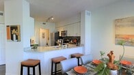 This well-appointed two-floor residence showcases a thoughtfully designed interior with modern finishes and a clean, contemporary aesthetic. The open-concept kitchen serves as a central highlight, featuring crisp white cabinetry that extends to the ceiling, providing generous storage. A mosaic tile backsplash adds a decorative focal point, complementing the granite countertops that wrap around the breakfast bar. The bar seating area, lined with upholstered stools, creates a natural transition to the dining space, making the layout ideal for both everyday living and entertaining. Stainless steel appliances, including a refrigerator and built-in microwave, complete the kitchen with a polished, functional feel. Track lighting illuminates the space efficiently, enhancing the bright, airy atmosphere created by the light-toned walls and cabinetry. The dining area adjacent to the kitchen offers a dedicated space for meals, furnished with a full dining table and coordinating chairs. The layout flows seamlessly across two floors, offering distinct zones for living and private quarters. Overall, the space presents a cohesive, modern design with attention to detail, practical functionality, and a welcoming ambiance suited for contemporary living.