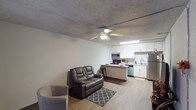 This single-floor unit offers a practical and efficient layout, making smart use of its space throughout. The open-concept design seamlessly connects the living area and kitchen, creating a cohesive and functional living environment. The living space is anchored by a ceiling fan with integrated lighting, set against a textured ceiling that adds subtle visual character. Light-toned laminate flooring runs throughout, contributing to a clean, modern aesthetic.

The kitchen is thoughtfully designed with white cabinetry providing ample storage, complemented by stainless steel appliances including a refrigerator, microwave, and range. A peninsula-style counter extends into the living area, offering additional workspace and casual seating with barstool access. The kitchen also features a stainless steel sink beneath under-cabinet lighting, adding both function and brightness to the cooking space.

The living area comfortably accommodates seating furniture and benefits from the open sightlines created by the shared layout. Neutral wall tones throughout the unit keep the space feeling light and versatile, allowing for a variety of furnishing and décor styles.
