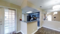 This single-floor residence offers a thoughtfully designed layout that balances functionality and style. The kitchen serves as a central highlight, featuring bold navy blue cabinetry that creates a striking contrast against the neutral warm-toned walls. A full suite of black appliances — including a refrigerator, gas range, and built-in microwave — are seamlessly integrated into the space. A tasteful tile backsplash adds texture and visual interest behind the range. The kitchen is topped with granite-style countertops and opens to the adjacent living area via a peninsula-style breakfast bar, promoting an open and connected feel throughout the home. Overhead, a recessed skylight panel provides additional illumination in the kitchen. The flooring transitions from rich, dark hardwood-style planks in the living area to lighter tile in the kitchen, clearly delineating each space while maintaining a cohesive aesthetic. A textured ceiling adds dimension throughout. Natural light filters in through multiple windows dressed with decorative curtains, enhancing the bright and welcoming ambiance. Passageways visible from the main area suggest additional rooms, rounding out this comfortable and well-equipped single-level home.