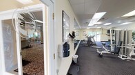 This single-floor amenity space offers a comprehensive set of shared facilities designed to support both active and social lifestyles. The fitness center is a standout feature, spanning a generously sized room with dark, cushioned flooring and large wall-to-wall mirrors that enhance the sense of space. It is well-equipped with a multi-station weight machine, cardio equipment including treadmills, ellipticals, and a stationary bike, along with exercise mats and a flat-screen TV. Bright recessed panel lighting and windows overlooking greenery create an energizing workout environment.

Adjacent to the fitness area, the lounge offers a warm, relaxed atmosphere anchored by a corner gas fireplace with a tile surround and a wall-mounted flat-screen TV above a sleek mantle. Rich, patterned carpeting and deep leather armchairs contribute to its inviting ambiance, while windows allow natural light to filter in.

The dining area features light wood tables and chairs set atop similarly patterned carpeting, with a glass door providing direct outdoor access and natural views. Nearby, a functional kitchen is outfitted with light maple cabinetry, dark countertops, a black electric range, and ample storage, serving as a practical support space for gatherings and events throughout the facility.