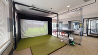 Located at 7145 Anderson Dr in Whitestown, Indiana, this single-floor game room is a well-appointed recreational space designed for social entertainment and active play. The room is finished with a two-tone gray and white wall treatment, recessed lighting, and durable carpeted flooring that ties the space together cohesively.

The centerpiece of the room is a fully enclosed indoor golf simulator powered by Foresight Sports technology, complete with a hitting mat, projection screen, and protective netting — offering an immersive experience for golfers of all skill levels. Adjacent to the simulator, a full-size shuffleboard table serves as another focal point for group activity, accompanied by red metal stools for casual seating.

A comfortable gray sectional sofa provides a relaxed lounge area, ideal for spectators or socializing between games. The space also includes a billiards area, wall-mounted flat-screen TV, and wooden bench seating along the perimeter. Decorative signage and wall art add character and a lively atmosphere throughout.

Large windows allow natural light to filter into the lounge area, while glass-panel doors connect the game room to an adjacent fitness center, enhancing the sense of openness. The overall ambiance is energetic yet comfortable, making this space a versatile hub for recreation.
