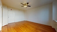 This single-floor unit offers a clean, practical layout with several notable design elements throughout. The main living area is spacious and open, showcasing warm-toned hardwood floors that run throughout, complemented by crisp white walls and white trim. A ceiling fan with an integrated light fixture is centrally mounted, providing both air circulation and ambient lighting. Two windows along one wall flood the room with natural light, and a built-in shelving unit beneath them adds a functional storage element. The entry door features classic paneled detailing, contributing a traditional touch to the otherwise simple aesthetic.

The kitchen is efficiently arranged, featuring warm wood cabinetry with upper and lower storage, granite-style countertops, a white electric range, and a full-size refrigerator. A window above the counter offers a cityscape view, adding a striking visual element to the cooking space. The floor transitions to light ceramic tile in this area, offering a practical surface.

The bathroom maintains a classic design with white square wall tiles framed by a bold cobalt blue tile border, adding a distinctive decorative detail. The space includes a full soaking tub, a pedestal sink, and a shower curtain rod, delivering a clean and functional layout overall.