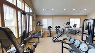 Located at 3025 Fountain Dr. in Beavercreek, Ohio, this single-floor fitness center is a comprehensively outfitted space designed to support a full range of workout routines. The room is defined by soaring vaulted ceilings accented by a ceiling fan and recessed lighting, creating an open and airy atmosphere. Neutral-toned walls and warm wood trim details lend the space a clean, welcoming aesthetic, while carpeted flooring provides comfort underfoot throughout the facility.

Natural light flows generously through multiple windows fitted with blinds, keeping the environment bright and inviting. The cardio section includes treadmills, upright and recumbent bikes, and an elliptical machine, offering a variety of options for cardiovascular training. Strength training is equally well-represented, with a cable crossover machine, a Nautilus multi-station unit, a power rack with an adjustable bench, and a dedicated dumbbell rack stocked with a full range of free weights and weight plates.

Additional equipment such as a stability ball, a wall-mounted flat-screen television, a large mirror, and a coat hook station further enhance the functionality and convenience of the space. French doors provide access to the fitness area, adding an architectural touch that complements the overall design. This facility is thoughtfully arranged to accommodate both individual training sessions and group use with ease.